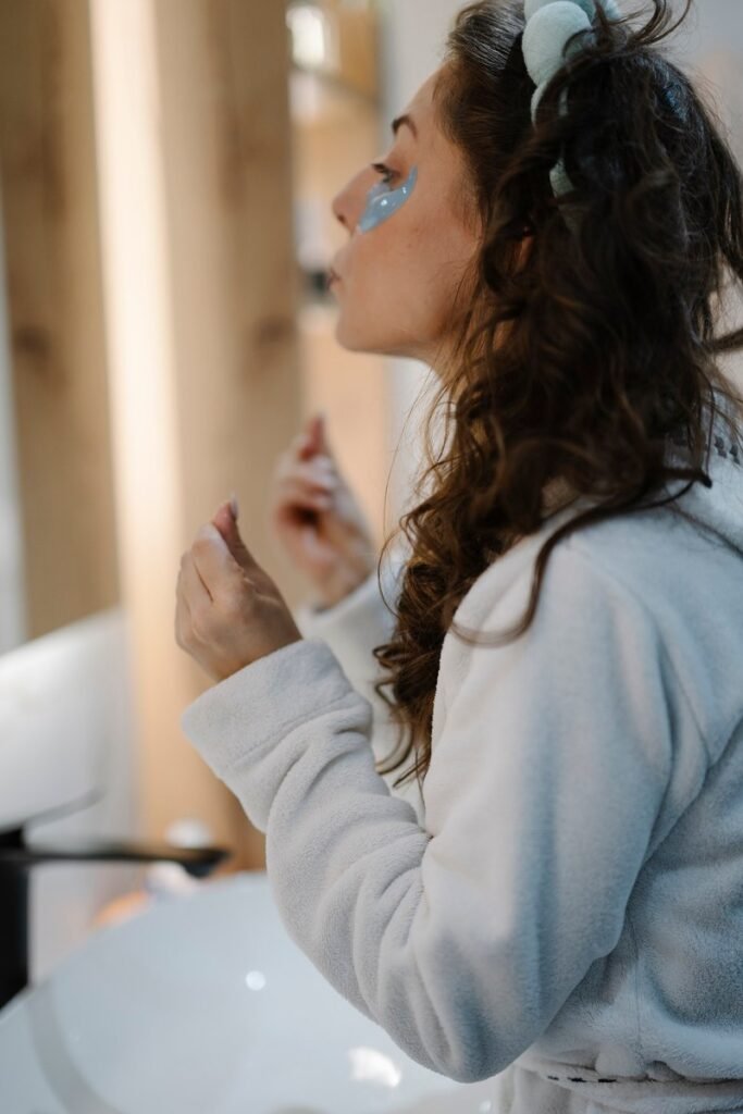 一名女性穿着浴袍、头戴发圈，在浴室镜前进行护肤，眼下贴有眼膜贴片，自然光线照入室内